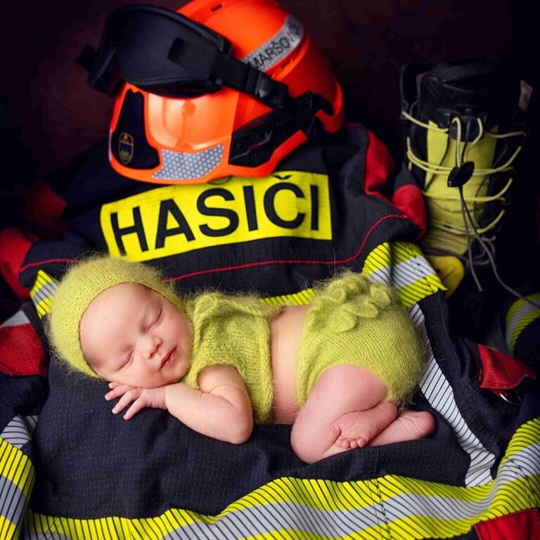 Hasičské téma (žluté): „Originální newborn focení Praha, miminko v pleteném žlutém setu spící u hasičské přilby a nápisu HASIČI.“