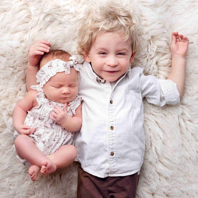 Dva sourozenci: „Focení dětí a miminek Praha, starší bratr ležící v objetí se svou novorozenou sestřičkou na světlém podkladu.“