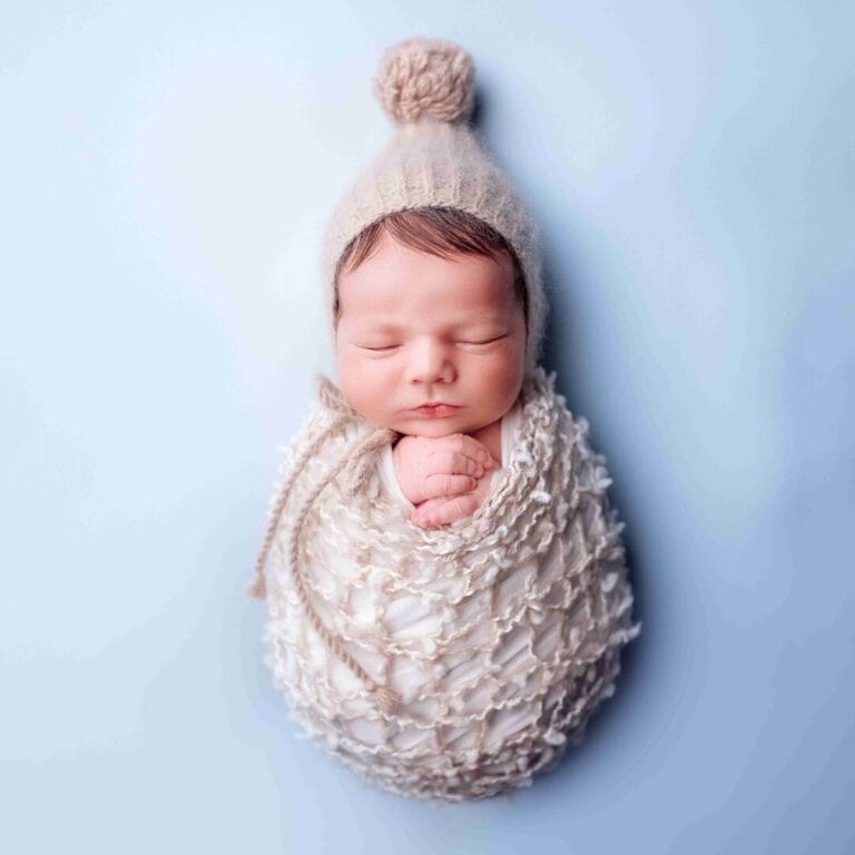 Detail spícího miminka v pleteném vaku s kulichem na modrém pozadí – newborn foto